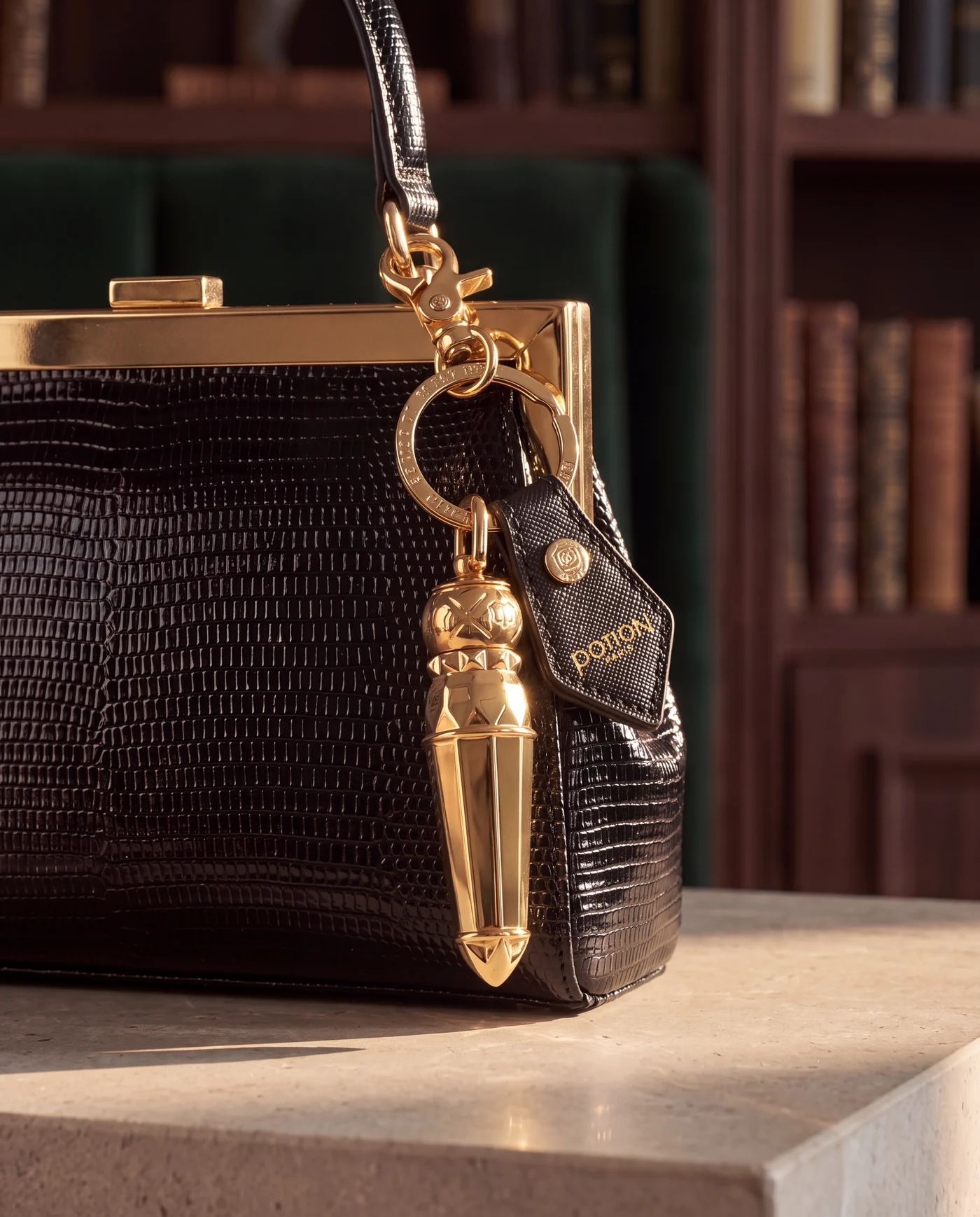 A gold vial pendant hanging from a dark lizard-skin handbag against a library backdrop.